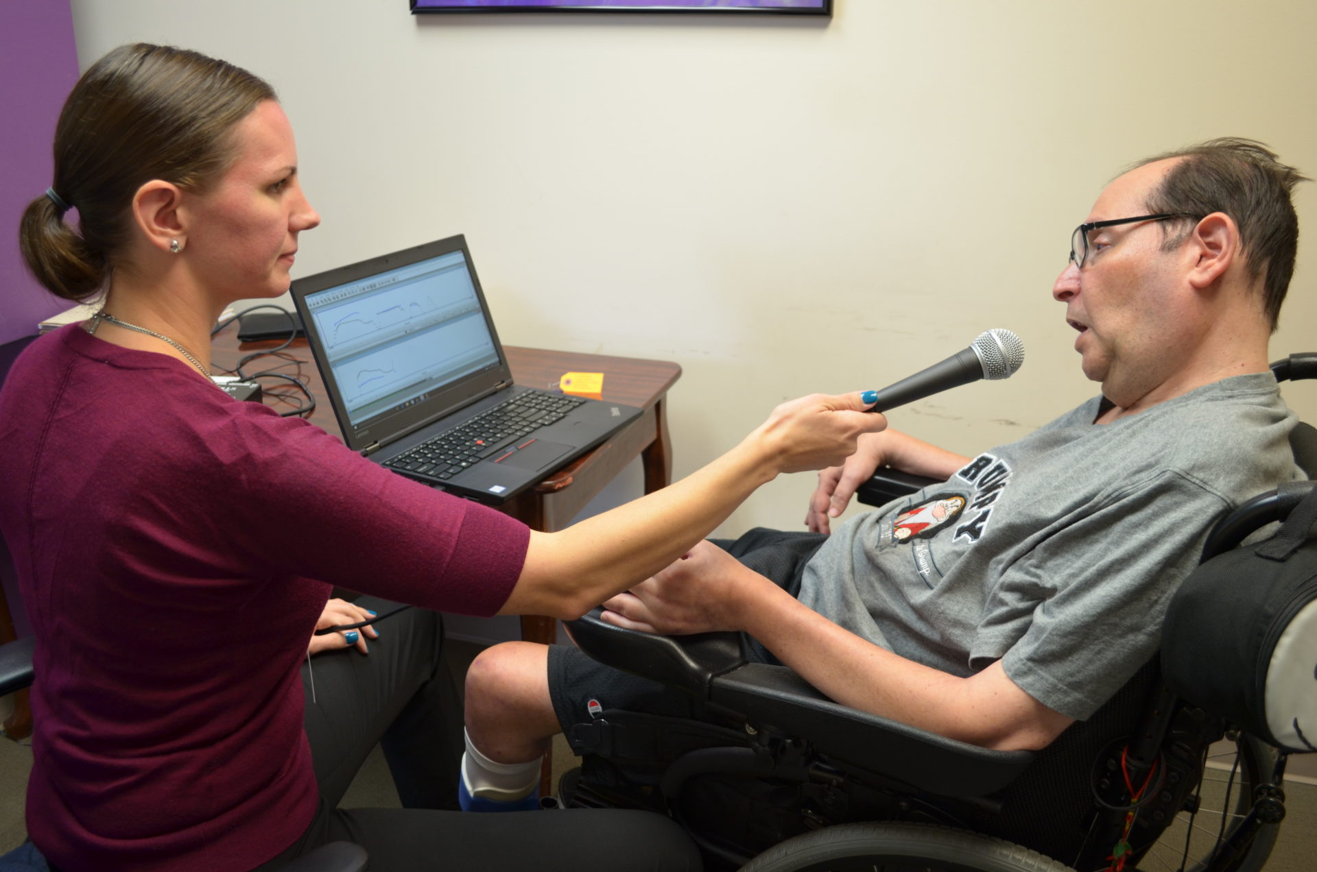 Bancroft NeuroRehab clinician holding a microphone up to a patients mouth while sitting by a desk with a laptop on it.