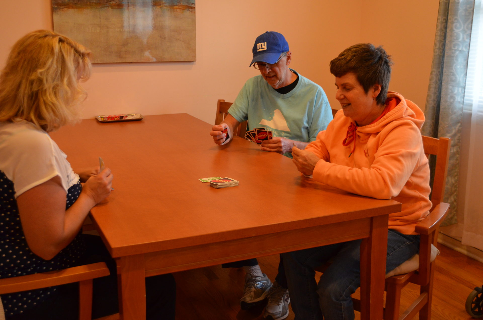 One older man and two older women playing Uno at a wooden table. Older man is wearing a New York Giants hat