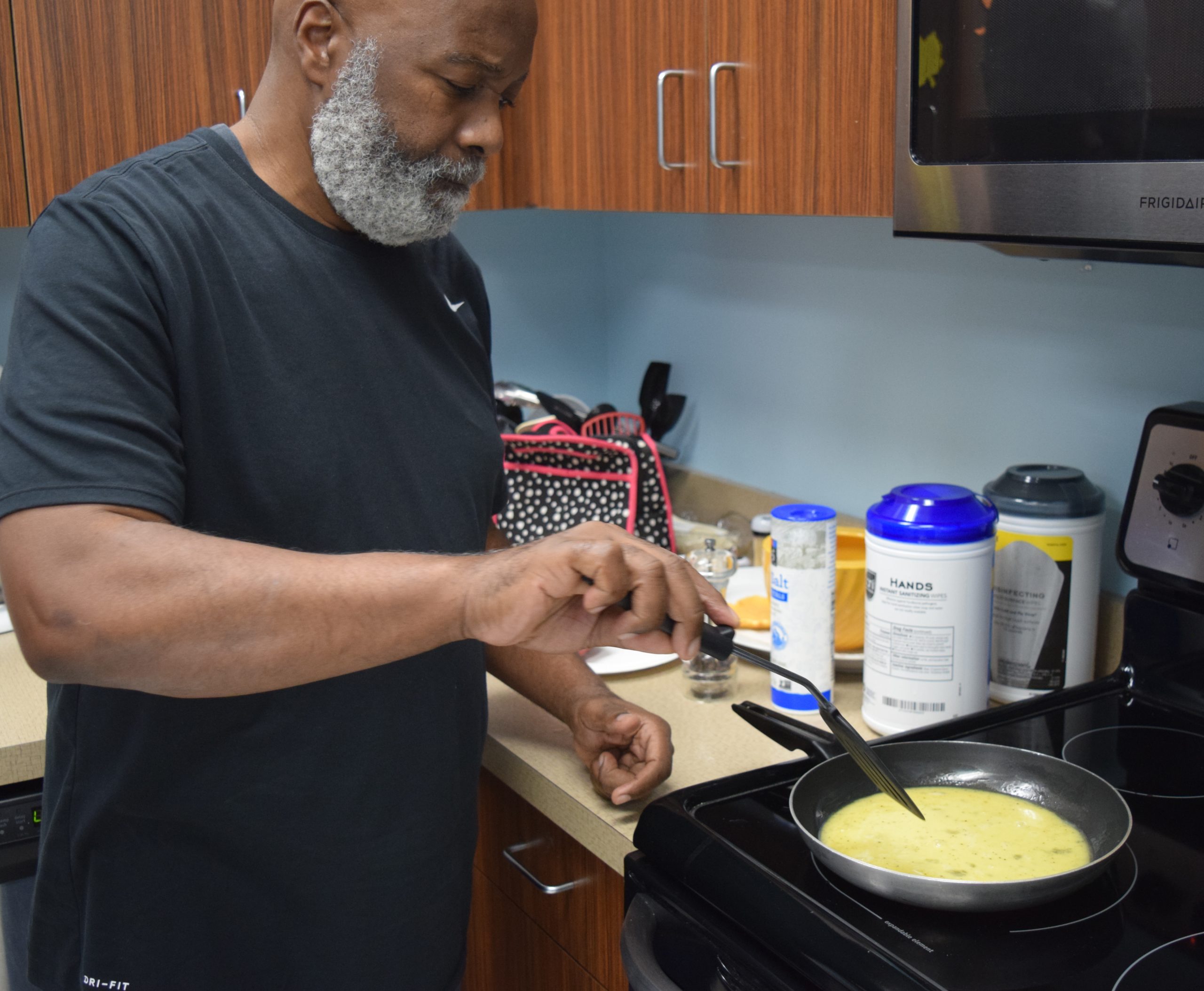 Man at a stove cooking eggs with a spatula in his right hand.