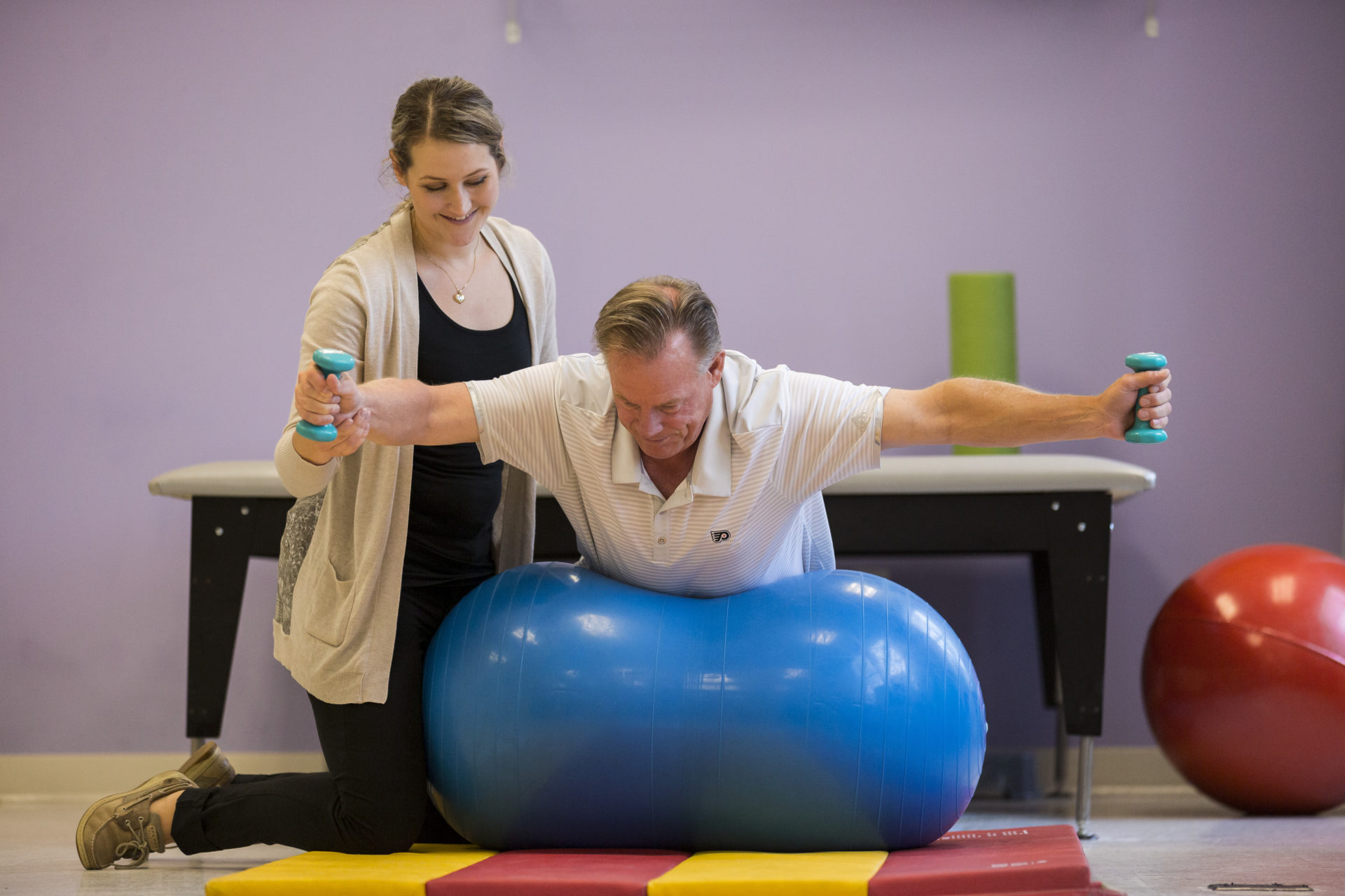 A Bancroft NeuroRehab clinician helping Brian Propp during a Physical Therapy Session. Brian is laying on an excercise ball while holding weights in each hand. His arms are spread out.
