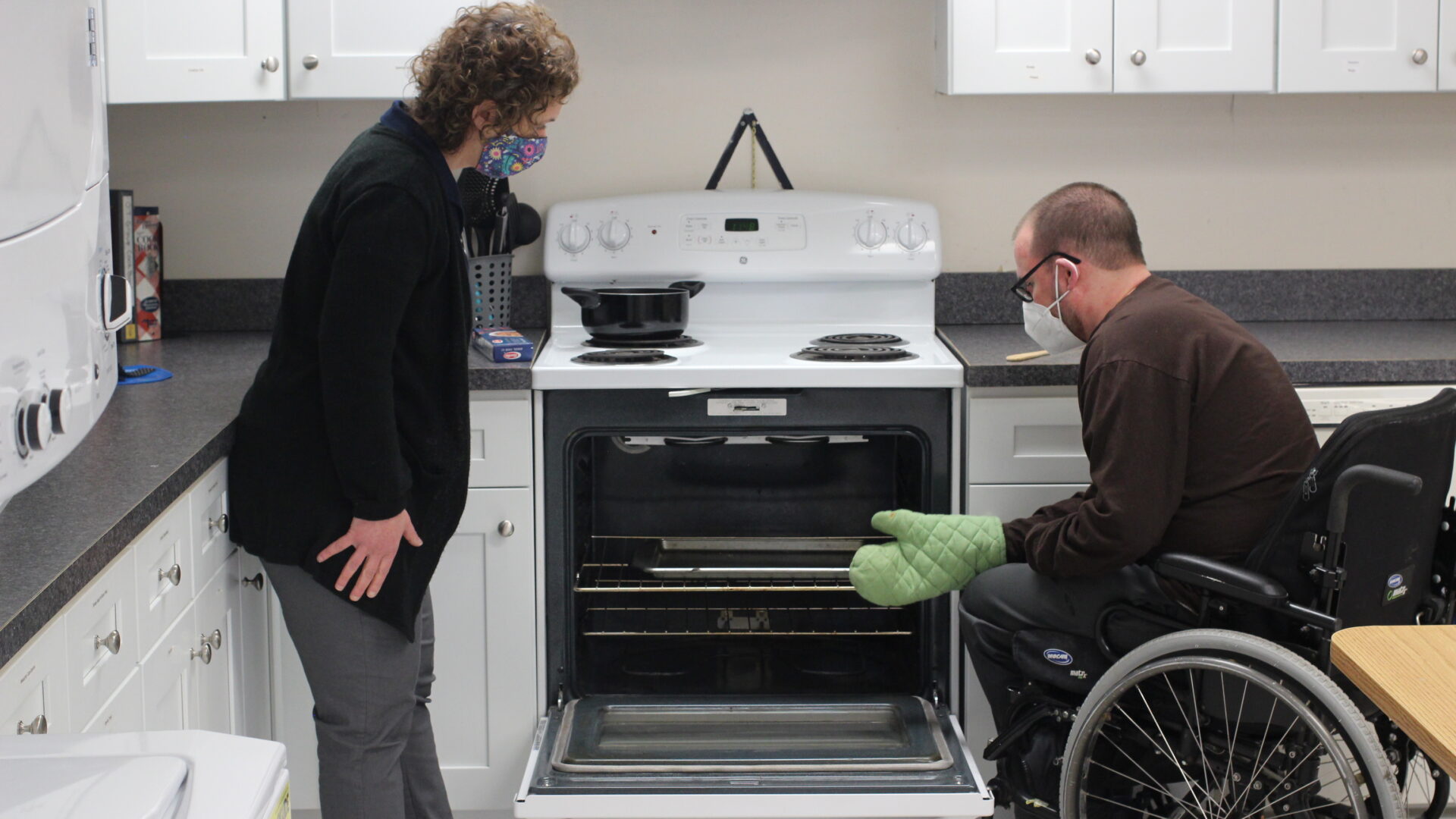 A Bancroft Neuro Rehab clinician and patient in a wheelchair are in a kitchen. They are both looking inside an open oven.